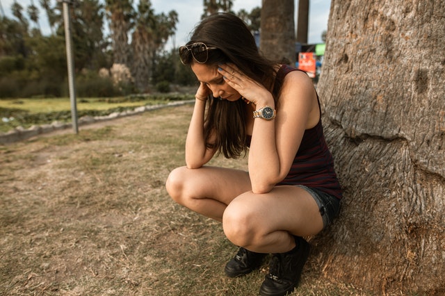 Women on trail sitting with her back against a tree and her hands on the temples of her head